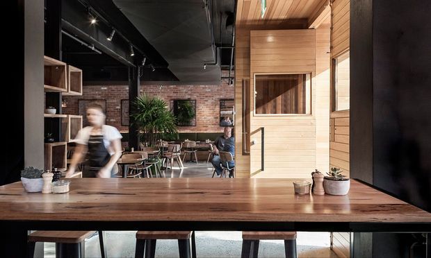 A warmly lit contemporary commercial café or hospitality interior viewed from a timber counter. The foreground features a substantial timber benchtop with stools, potted plants, and a large dark display or screen to the right. Beyond it is an open dining area with tables and chairs, exposed red-brick walls, black-painted ceiling services and track lighting, timber-framed shelving, and a timber-clad architectural volume with a small interior window. People are present but not the focus.