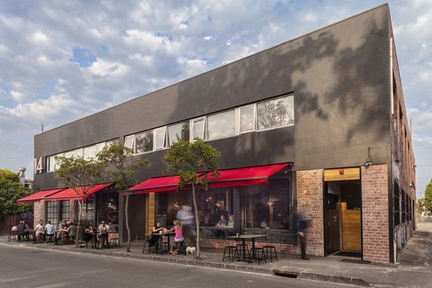 Street-level photograph of a two-storey urban commercial building, likely a café or restaurant, with a long dark rendered upper facade, exposed brick side walls, extensive ground-floor glazing, red fabric awnings, outdoor dining tables, and a prominent timber-framed entrance. The building has a simple rectangular form and a flat roof, set directly along a paved street.