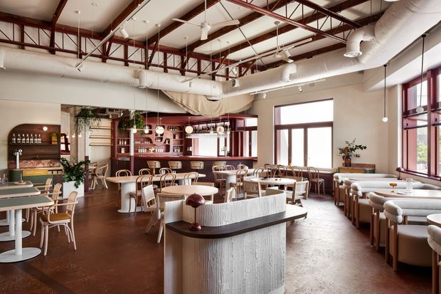 A spacious industrial-style café or restaurant interior with a high exposed ceiling, red-painted steel roof trusses, visible white mechanical ductwork, and large burgundy-framed windows. The space contains a central fluted service counter, a timber-backed bar with open shelving, numerous small round dining tables, upholstered banquette seating, bentwood-style chairs, pendant lighting, and indoor greenery. The palette combines warm timber and terracotta-brown flooring with cream walls, white ceilings and ducts, and dark red structural and window accents.