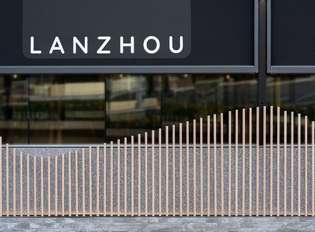Close-up exterior view of a contemporary commercial building facade. A dark charcoal sign reading “LANZHOU” sits above reflective glazing, while a textured speckled wall is screened by closely spaced pale vertical metal rods forming a gentle wave profile. The lower edge shows a grey masonry or concrete base.
