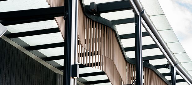 Close-up architectural view of a contemporary facade or covered exterior passage, featuring dark steel framing, translucent roof panels, curved black trim, and a suspended screen of closely spaced vertical timber or wood-toned battens. The composition emphasizes layered structure, rhythm, and modern material detailing.
