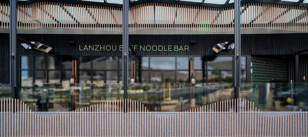 Exterior view of a contemporary commercial noodle bar facade. The frontage uses dark vertical cladding, extensive glazed shopfronts, and prominent rhythmic vertical slatted screening across the upper and lower sections. The sign reads “LANZHOU BEEF NOODLE BAR.”