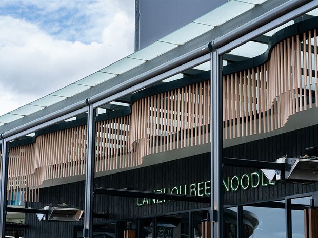 Close-up exterior view of a contemporary commercial building facade. The dark vertically clad lower level features large glazing and a partially visible illuminated sign, while the upper level is screened by repeating pale timber-toned vertical slats with curved sculptural sections. A projecting translucent canopy and exposed dark metal drainage pipes run along the roof edge. Cloudy sky is visible above.