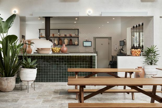 A bright contemporary café or hospitality interior with a green tiled service counter, open black shelving displaying ceramics and bottles, white tiled walls, timber picnic-style dining tables and benches, indoor plants, and polished concrete or stone-look flooring. Pendant lights and concealed lighting create a clean, welcoming atmosphere.
