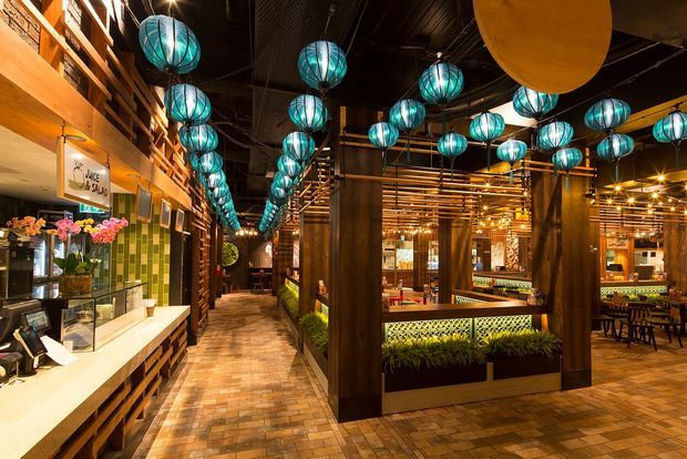 A richly detailed hospitality interior, likely a restaurant or bar, with a long entry and service counter on the left, exposed dark ceiling, extensive timber wall and ceiling detailing, brick-pattern flooring, green tiled feature wall, and numerous turquoise spherical pendant lights. Dining tables, banquette seating, planting, and warm ambient lighting extend through the open dining area.