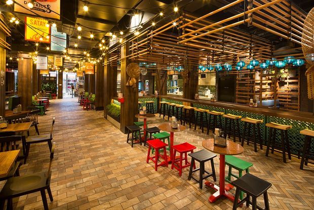 A warmly lit, bustling restaurant or bar interior with a long tiled floor, rows of small tables and brightly coloured stools, a timber bar counter, and extensive horizontal timber slat screening and ceiling detailing. String lights, pendant fixtures, patterned tile surfaces, signage, and decorative blue lanterns create an eclectic, lively hospitality atmosphere.