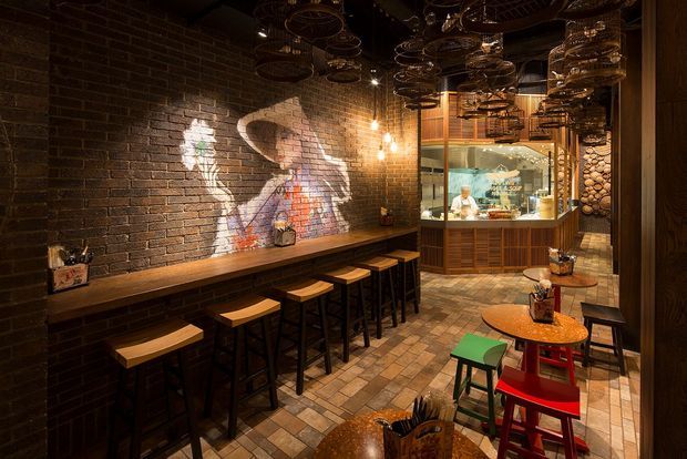 A dimly lit industrial-style restaurant or bar interior with exposed dark brick walls, timber counter seating, black metal stools, patterned tile flooring, and a compact service counter at the rear. Hanging metal cookware or basket-like fixtures and warm pendant lighting create a dramatic, rustic atmosphere, while colourful stools and projected artwork add visual accents.