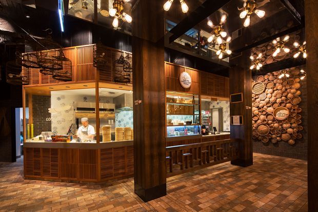 A warmly lit restaurant or café interior with a strong industrial-rustic character. Dark timber columns and ceiling beams frame a food preparation counter and bar, while exposed brick paving, timber slatted cabinetry, glass display shelving, and decorative pendant fixtures create a rich, atmospheric fit-out. A feature wall displays circular timber pieces, and an upper mezzanine with metal railings is visible.