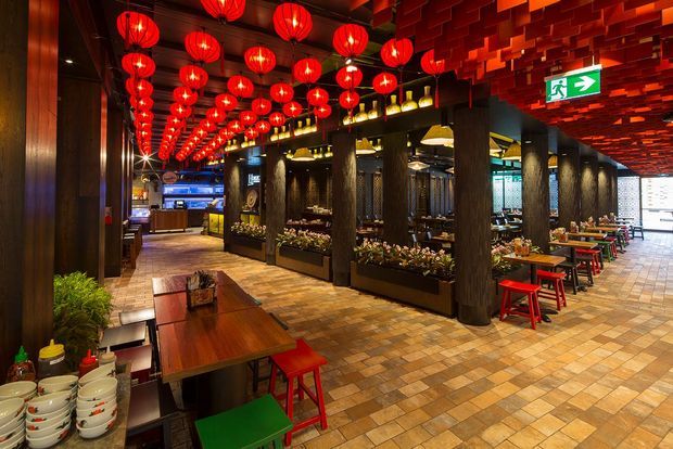 A spacious, richly decorated restaurant interior with dark timber columns and walls, an exposed patterned ceiling, rows of glowing red lanterns, tiled flooring, communal timber tables, and colourful red and green stools. A bar and dining area extend into the background, with illuminated exit signage and planter boxes integrated into the seating partitions.