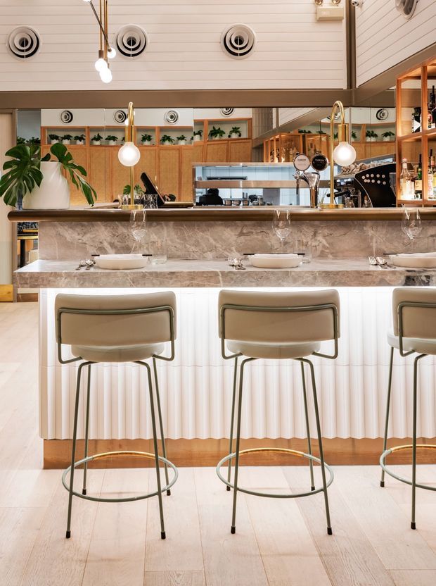 A refined hospitality bar or restaurant interior viewed from the dining side. A long marble-faced counter with integrated fluted white detailing is paired with three upholstered bar stools. The space features timber cabinetry and shelving, mirrored wall panels, brass-toned details, globe pendant lights, indoor greenery, and a white horizontal-panel ceiling with exposed circular ventilation grilles.