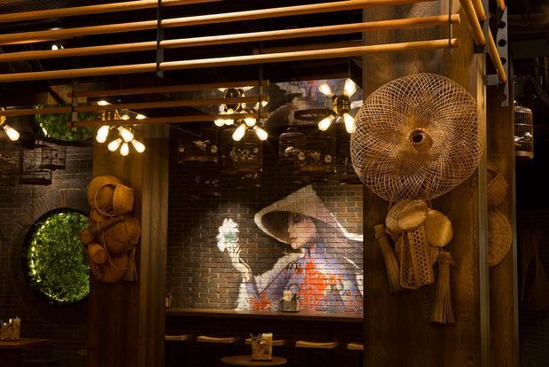 A dark, atmospheric interior featuring exposed red-brown brick walls, timber posts and overhead timber slats, warm pendant lighting, and extensive woven rattan and wicker decor. A large circular woven wall feature and hanging sculptural pieces frame a mural-like artwork above built-in bench seating, creating an eclectic, rustic restaurant or hospitality setting.
