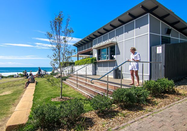 A modern single-storey building beside a beachfront promenade, featuring grey panelled walls, extensive glazing, a deep projecting roof with exposed structural framing, timber steps, and an accessible ramp with metal handrails. Landscaped planting and a paved walkway surround the building, with the ocean and beach visible in the background.