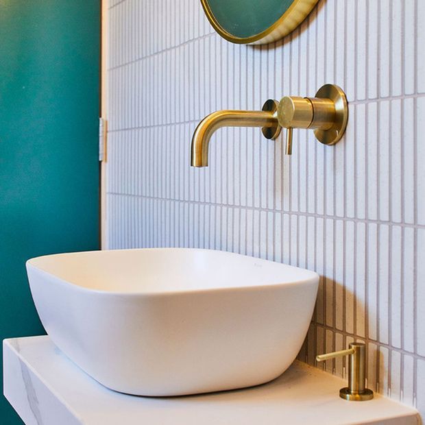 Close-up of a contemporary bathroom vanity featuring a white vessel basin on a white benchtop, a wall-mounted brushed-gold tap, and a matching gold basin mixer. The wall is finished in narrow white vertical tiles, with a bold teal painted wall and a partial round mirror adding colour and contrast.
