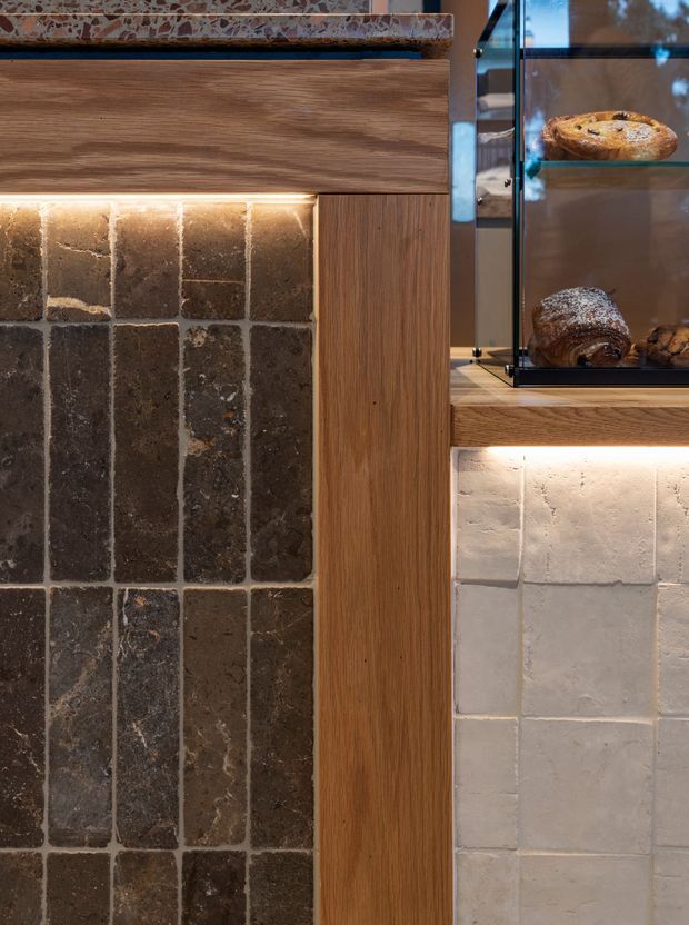 Close-up detail of a refined food-service or kitchen fit-out featuring dark rustic stone or ceramic tile, pale handmade-style wall tiles, vertical timber joinery, a speckled stone shelf, and warm integrated strip lighting. A glass display cabinet with baked goods is visible on the right.