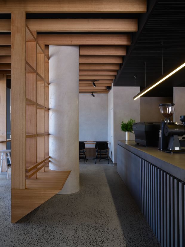 A refined contemporary café interior with a long counter and espresso machine on the right, a dark vertically fluted counter front, exposed black ceiling services, and a striking linear pendant light. Warm timber shelving, integrated bench seating, and a timber batten ceiling surround a rounded concrete column. Small tables and stools sit in the background, while the polished speckled concrete floor reinforces the modern hospitality setting.