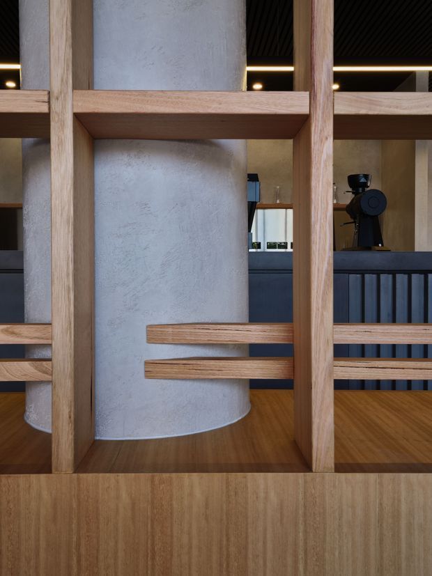 Interior architectural detail featuring a warm timber screen and shelving framework in front of a large textured cylindrical plaster/concrete column. A dark-toned counter with coffee equipment is visible behind the screen, with timber flooring, dark ceiling slats, and integrated linear lighting creating a refined hospitality or café-like atmosphere.