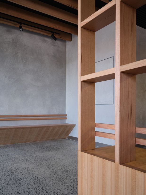 A contemporary interior featuring a sculptural timber built-in divider and bench seating. The open shelving structure uses warm natural wood, separating the foreground from a grey plastered wall and a recessed seating area. The room has a dark ceiling with exposed timber elements and track lighting, while the floor is finished in grey terrazzo or polished concrete with visible aggregate.