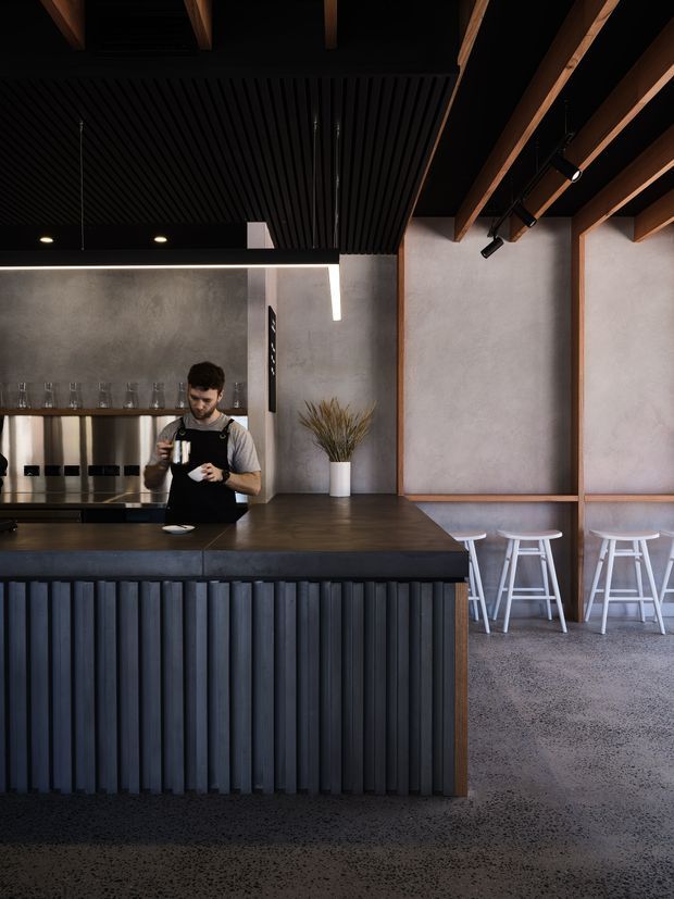 A dark, refined commercial bar or café interior featuring a large black island counter with vertically ribbed detailing, a dark timber benchtop, integrated shelving and glassware, and a staff member preparing a drink. The space combines polished concrete flooring, grey plaster walls, exposed timber ceiling beams, black slatted ceiling panels, track lighting, and white bar stools along a window-side counter. The overall atmosphere is moody, contemporary, and industrial-inspired.