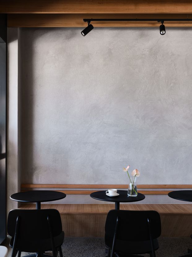 A refined café or commercial dining space with a long textured light-grey plaster feature wall, warm timber ceiling and wall panelling, black round pedestal tables, and black chairs. Minimal black track spotlights illuminate the wall, while a small central table holds a coffee cup and a delicate flower arrangement. The image emphasizes the material palette and built-in banquette-like timber ledge rather than the full room.