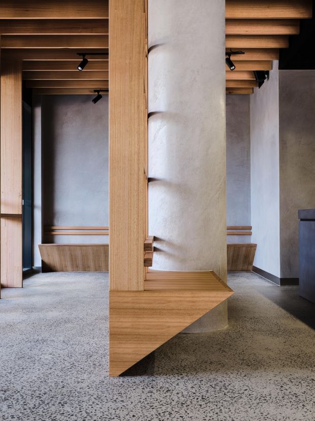 A close architectural interior view centred on a large cylindrical rendered column integrated with a sculptural timber built-in bench or joinery element. The space features exposed timber ceiling battens, warm timber wall framing, pale textured plaster walls, black track lighting, and a polished aggregate or terrazzo-like concrete floor. The composition emphasises custom detailing, layered structure, and a warm, refined material palette.