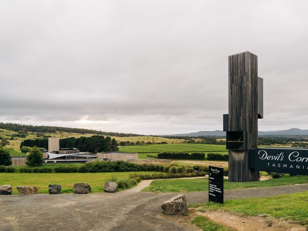A landscaped rural vineyard entrance in Tasmania, featuring a tall dark vertical architectural sign structure, a low modern winery building, gravel approach, large rocks, and expansive green fields beneath an overcast sky. The scene is primarily an exterior view of the winery grounds and building facade.