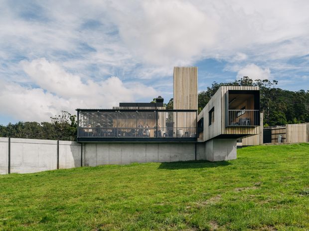 A contemporary multi-level residential house set into a gently sloping grassy site. The building features exposed concrete retaining and foundation walls, dark-framed glazing, timber-look vertical cladding, projecting box-like volumes, a covered outdoor room or terrace, and a tall vertical tower element. Dense trees form the background, while the dramatic roofline and cantilevered upper volume create a sculptural modern facade.