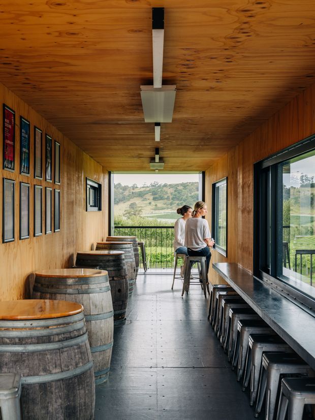 A narrow, wood-lined tasting room or bar interior with barrel tables along one wall and a long dark counter with stools beneath large windows on the other. Framed displays decorate the timber wall, while an open glazed end provides views of rolling countryside. Two people sit at the far end, and simple linear pendant lighting runs along the timber ceiling.