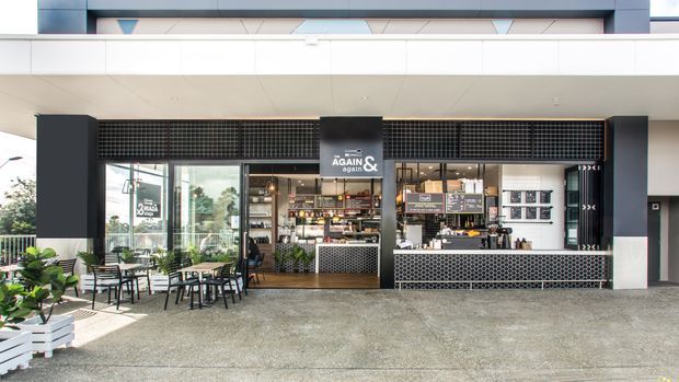 A modern café or food-and-beverage kiosk set within a covered commercial arcade. The frontage features black framing, extensive glazing, patterned black tile screens, open service counters, shelving, and outdoor café seating with planters. The palette is predominantly black, white, grey, and natural timber tones.