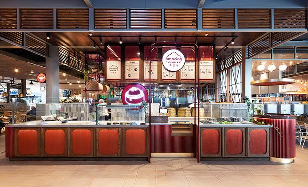 A vibrant commercial food-service interior featuring a central takeaway counter and branded kiosk, surrounded by open kitchen and dining areas. The fit-out combines dark metal framing, warm timber slatted screens and ceilings, red ribbed and upholstered counter panels, tiled flooring, pendant globe lights, and prominent illuminated signage.