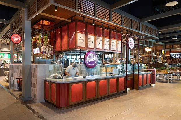 Interior view of a contemporary commercial food-service venue, centred on a compact restaurant or takeaway counter with glossy red upholstered or panelled cabinetry, illuminated menu boards, glass display and preparation areas, and overhead timber louvres. The wider dining area contains tables and chairs, with exposed dark ceiling services, pendant lighting, tiled flooring, and industrial-style metal framing. The composition is bright and polished with strong red and timber accents.