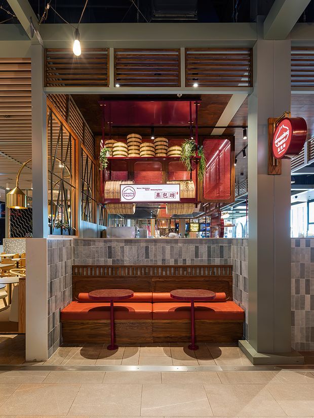 A compact, richly detailed restaurant booth in a contemporary commercial interior. The built-in booth has vivid orange-red upholstered bench seating, dark timber panelled joinery, small round tables, and a grey mixed-tile half wall. Above it is a decorative timber-louvred and glazed service/display area with hanging plants, stacked bamboo steamers, red illuminated surfaces, and Asian-inspired signage. Exposed dark ceiling services, structural beams, pendant lighting, and views into the broader food-hall environment reinforce the restaurant setting.