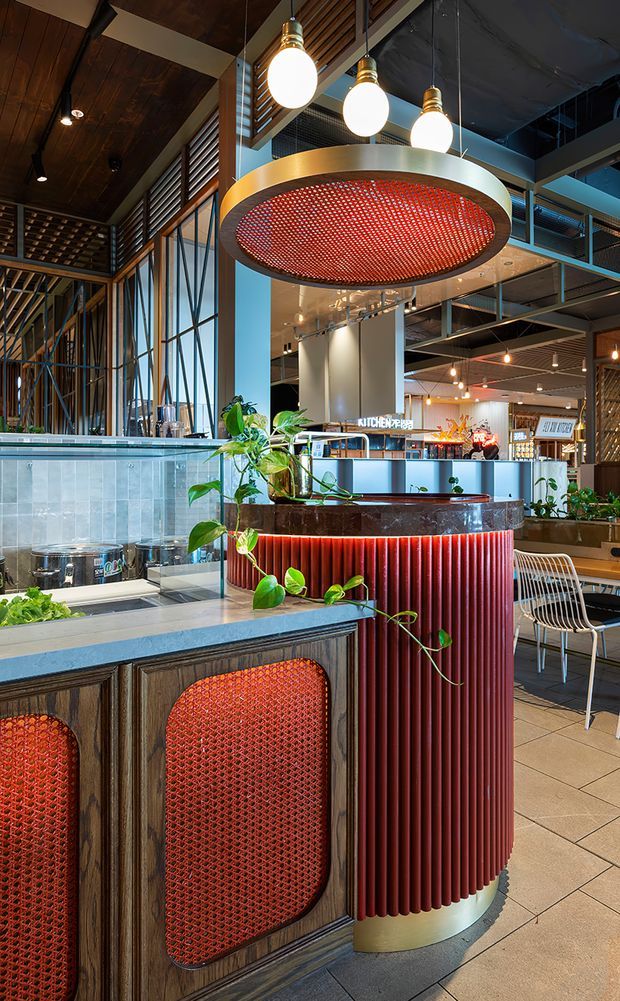 A richly detailed commercial hospitality interior, likely a restaurant or food hall, featuring a curved service counter with dark timber cabinetry, red ribbed cladding, and red perforated inset panels. A pale stone or solid-surface countertop, tiled flooring, tall glazed windows, timber ceiling slats, exposed services, and a large circular perforated feature pendant dominate the space. Warm globe pendant lights and abundant trailing plants add contrast to the industrial structural framework and mezzanine-like upper level.