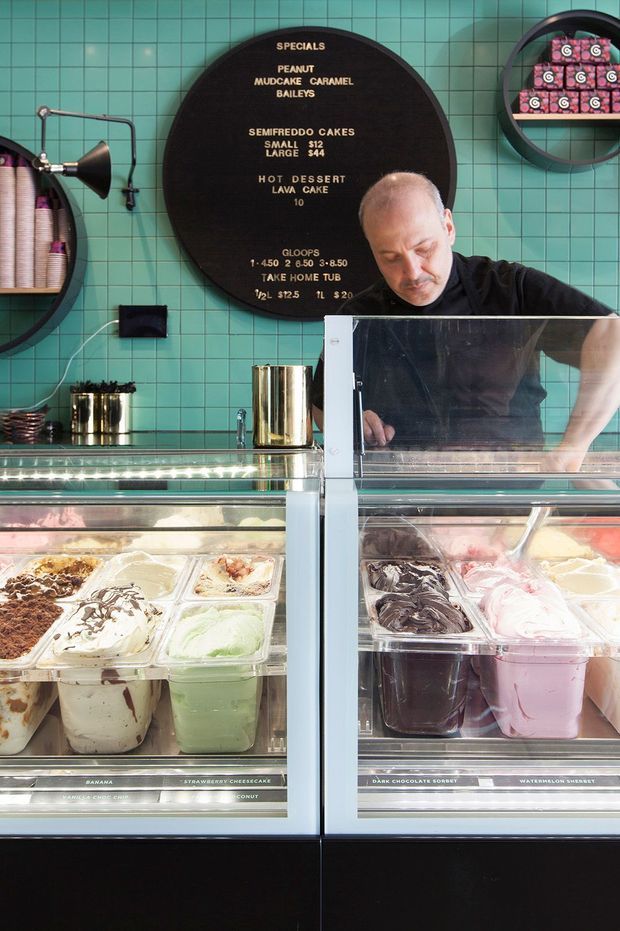 A brightly lit gelato or dessert shop counter with glass-front refrigerated display cases filled with multiple flavours of gelato and sorbet. A staff member stands behind the counter. The space features a turquoise green tiled wall, black circular menu board with gold lettering, black shelving, a wall-mounted industrial-style lamp, stainless-steel canisters, and dark counter bases. The image is a close, frontal view of a commercial food-service interior.
