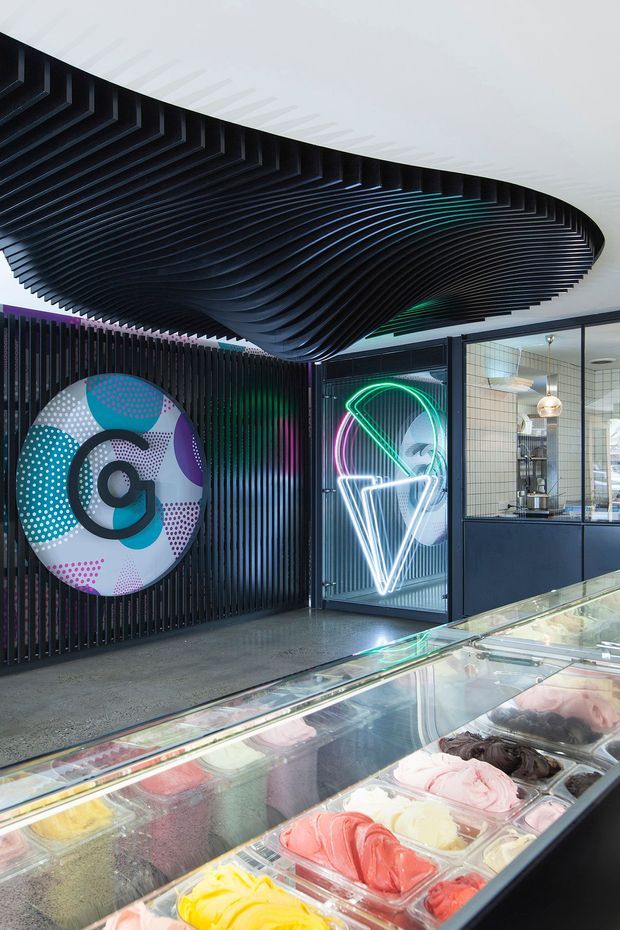 A vibrant contemporary gelato shop interior with a long glass-fronted refrigerated display counter filled with colourful gelato. The space features dark ribbed wall panels, a dramatic sculptural black wave-like ceiling installation, vivid neon geometric signage, and graphic circular wall art. Glazed partitions reveal additional commercial preparation or service areas.