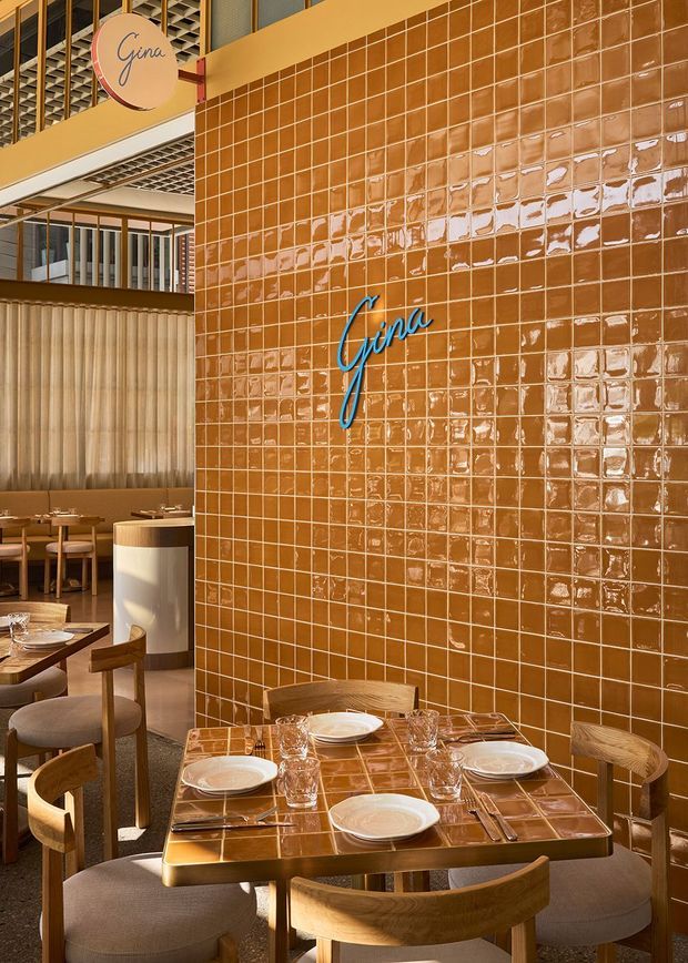 Interior view of a contemporary restaurant dining area centred on a glossy terracotta-brown tiled feature wall with illuminated cursive “Gina” signage. Set dining tables with timber chairs and upholstered seating occupy the foreground and left side, while sheer curtains, a curved service counter, dark timber framing, and an open patterned ceiling add depth. The palette is warm and refined, with strong tactile tile reflections and layered architectural detailing.