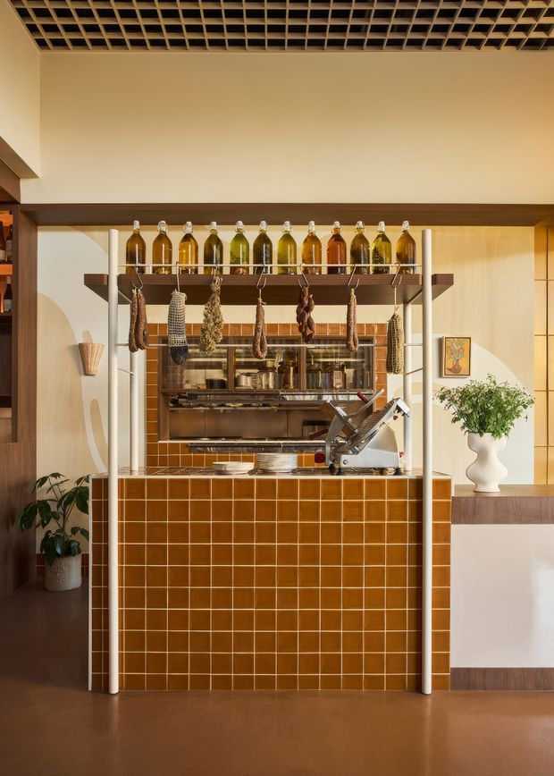 A warm, richly detailed hospitality-style interior featuring a compact tiled bar or serving counter with a commercial meat slicer, hanging cured meats and herbs, and rows of coloured bottles displayed on an overhead timber-and-metal rack. The space combines glossy terracotta-brown square wall tiles, timber shelving and trim, cream plaster walls, and a decorative exposed grid ceiling. Plants, ceramics, and artwork add character to the surrounding retail or food-service setting.