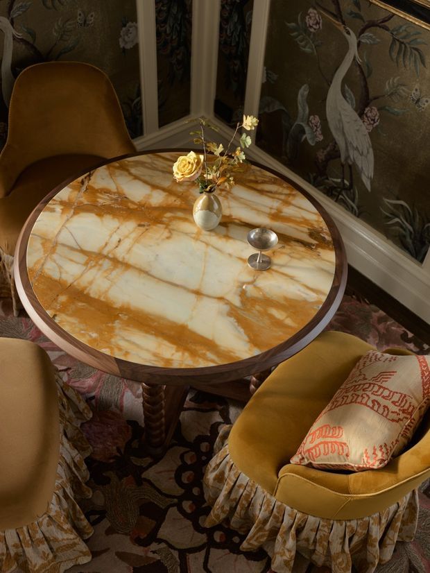 A richly styled, intimate seating area centered around a round table with a dramatic veined marble top and dark timber edge. Mustard velvet upholstered chairs with gathered fabric skirts surround the table, which holds a small vase of flowers and a silver decorative object. Dark botanical wallpaper with cranes, birds, and foliage forms the backdrop, while a patterned floral rug and embroidered cushion reinforce the ornate, layered aesthetic.