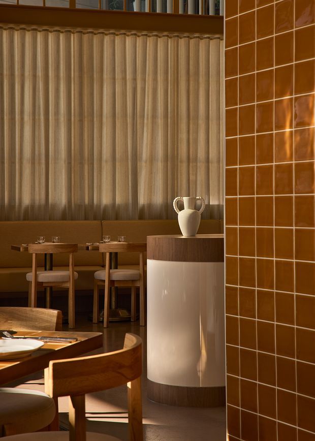 A warmly lit restaurant interior with a refined dining area. Built-in brown upholstered banquette seating lines the rear wall beneath tall sheer curtains, accompanied by small timber tables and chairs. In the foreground is a dining table, while a curved cylindrical timber-and-cream display counter with a ceramic vase separates the seating areas. A prominent glossy terracotta-brown tiled wall occupies the right side, and dark timber trim and flooring create a rich, intimate atmosphere.