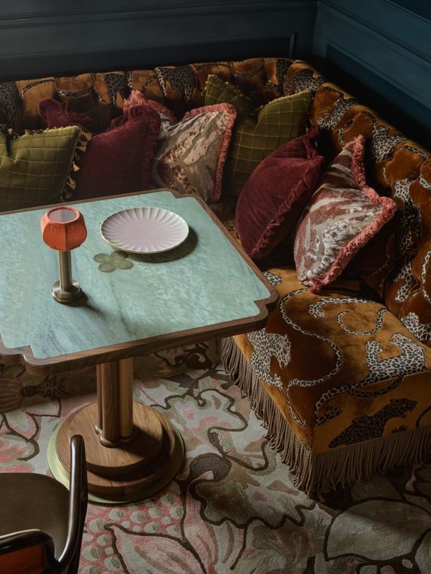 A richly styled, moody seating nook featuring a built-in or corner banquette upholstered in a bold brown animal-print and chain-patterned textile, layered with velvet and fringed cushions in burgundy, olive, pink, and patterned tones. A pale turquoise stone-look tabletop with a sculptural pedestal base sits in front, accompanied by a small orange table lamp and decorative plate. Dark blue-green wall panelling, an ornate patterned rug, and warm low lighting create an eclectic, luxurious atmosphere.
