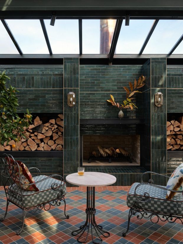 A stylish enclosed outdoor lounge or sunroom centered on a built-in fireplace with dark green glazed tile walls, integrated firewood storage, and a transparent pitched roof with dark structural framing. Two ornate wrought-iron lounge chairs with patterned cushions face a small round table. The floor features a colourful geometric tile pattern, with potted foliage and decorative objects adding warmth. The space has strong indoor-outdoor character and abundant natural light.