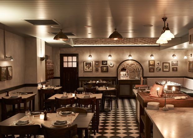 A warmly lit, intimate restaurant dining room with numerous dark timber tables and chairs, white table settings, framed artwork, wall sconces, pendant lights, and a striking black-and-white checkerboard floor. A long timber service counter occupies the right side, while an arched exposed-brick alcove and dark timber doorway provide architectural character.