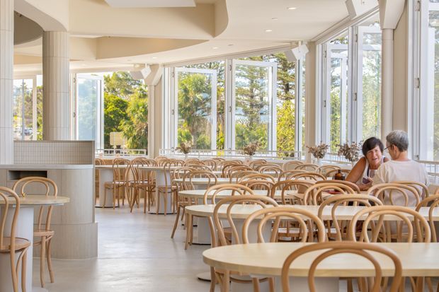 Bright, spacious contemporary café or restaurant interior with extensive floor-to-ceiling glazing, pale tiled columns, a light neutral ceiling and floor, and numerous light timber tables and bentwood chairs. Diners are seated near the windows, with leafy outdoor scenery visible beyond the glass.