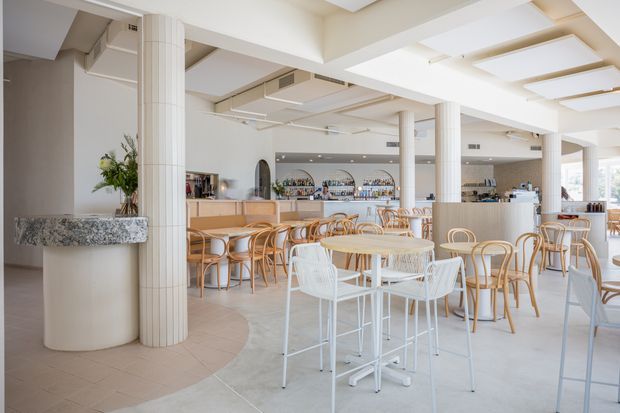 Bright, spacious contemporary hospitality interior resembling a café or restaurant, with a prominent curved bar, extensive bottle display, marble-topped feature column, fluted columns, and extensive white ceiling beams. The room is furnished with light timber and white dining tables and curved bentwood chairs, creating an airy, refined atmosphere.