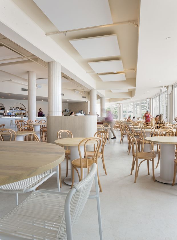 Bright, spacious contemporary café or restaurant interior with extensive glazing along one side, pale terrazzo or concrete-look flooring, white structural columns and ceiling, exposed services, and a large number of light timber and cane-style dining tables and chairs. A bar and service counter occupy the rear, while diffuse daylight creates an airy, welcoming atmosphere.