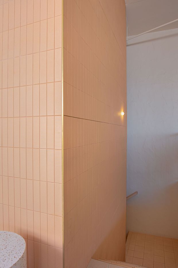 Close-up architectural interior detail of a warm peach and terracotta tiled bathroom or wet-area enclosure. Fine vertically textured rectangular tiles cover a curved or angled wall, with matching tiled flooring and a white textured plaster wall beside it. A small warm wall light creates a subtle glow, and the edge of a terrazzo-like curved vanity or basin is visible in the lower left.