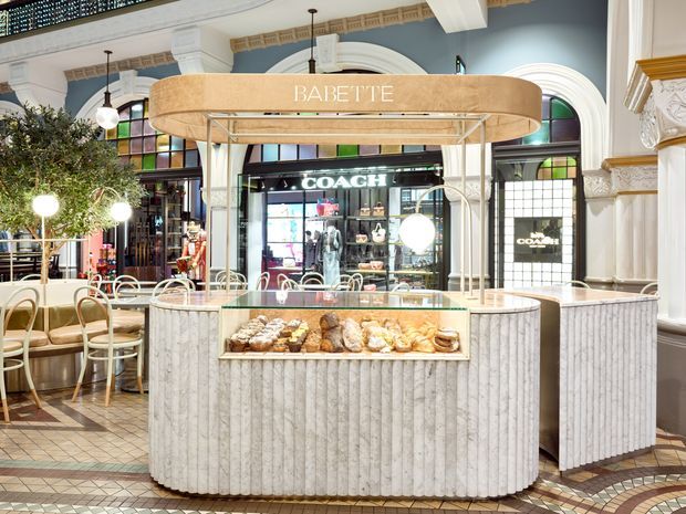A refined bakery or café kiosk named Babette in a grand heritage-style commercial interior. The central curved counter features fluted light stone cladding, a glass pastry display, and a rounded timber-toned overhead canopy with integrated lighting. Surrounding details include ornate white columns, arched stained-glass and glazed openings, decorative cornices, patterned tiled flooring, café seating, and a visible Coach storefront in the background.