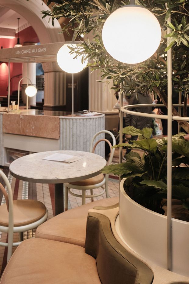 A warmly lit contemporary café interior with round marble-topped tables, curved upholstered seating, bentwood-style chairs, ribbed cylindrical counters, and abundant integrated greenery. Large glowing spherical pendant lights hang among tree branches, while a visible sign reads “UN CAFE AU...”. The space features arched openings, fluted architectural surfaces, and a sophisticated earthy palette of terracotta, tan, cream, and muted green.