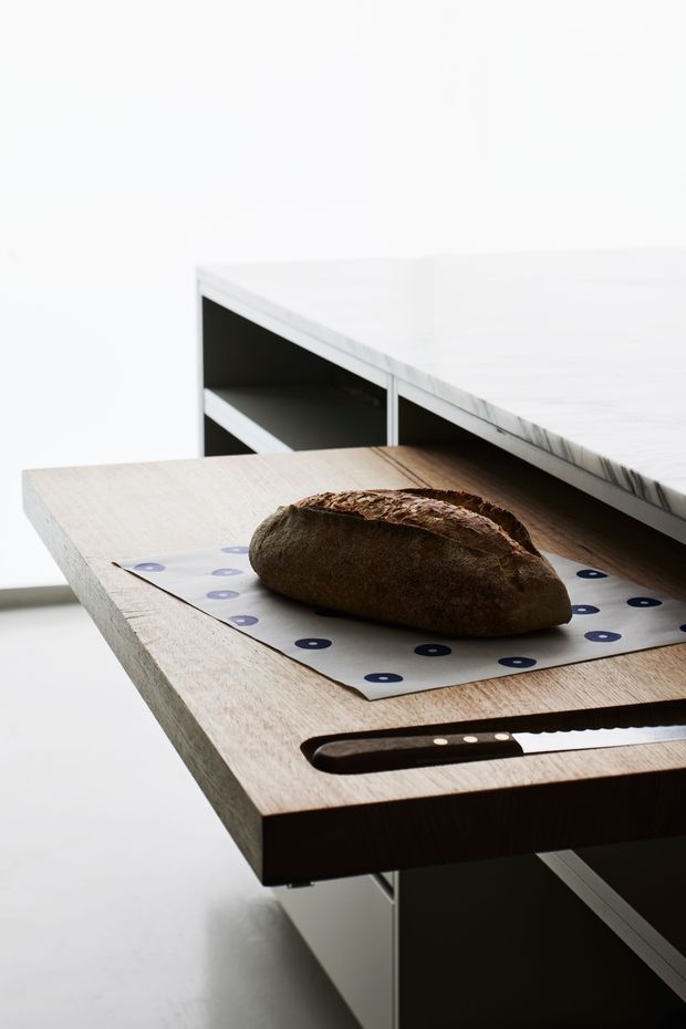 Close-up of a minimalist kitchen island or work surface featuring a thick light timber cutting board with a loaf of bread and bread knife. Dark open shelving and cabinetry sit beneath a pale white stone benchtop in the background, creating a clean, high-contrast contemporary kitchen composition.