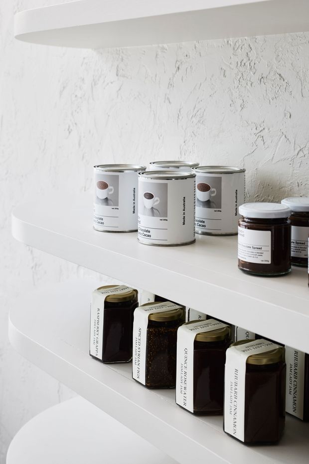 Close-up of white built-in shelving with thick rounded-front shelves against a heavily textured white plaster wall. The shelves display neatly arranged tins, jars, and glass containers, creating a bright, monochromatic product-display scene.