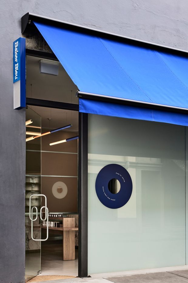 Street-level view of a contemporary retail storefront with a vivid blue retractable awning, dark metal framing, glazed entrance, and a vertical Baker’s Delight sign. The interior is partially visible through the open glass door and storefront, showing display shelving, counters, warm linear lighting, and polished finishes.